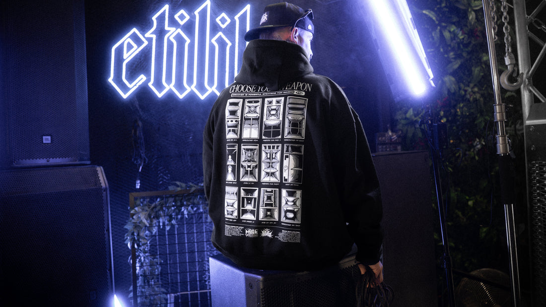 Hoodie noir Etilik « Choose your weapon » porté de dos devant l'enseigne néon, entre les enceintes